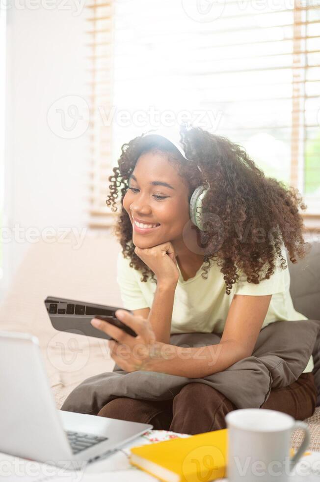 An attractive Black woman wearing headphones is enjoying watching a movie on her phone on the couch. photo
