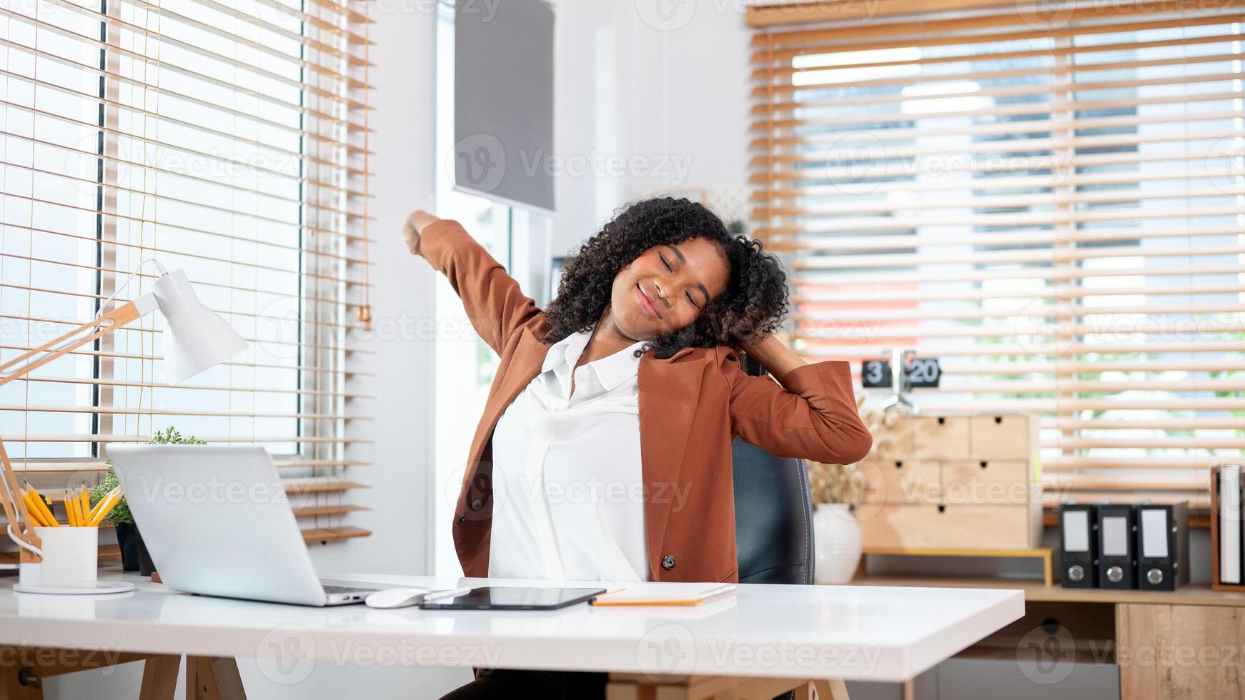 A positive Black businesswoman is stretching her arms and body at her desk in the office. photo