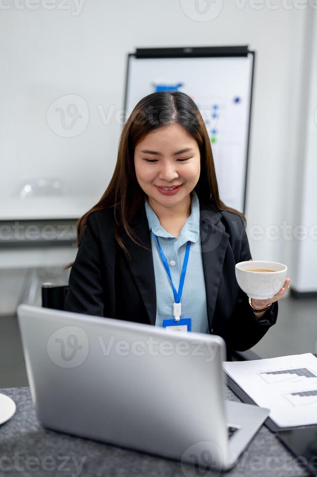A happy Asian businesswoman is having coffee while working on her laptop computer at her desk. photo