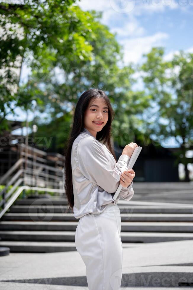 A beautiful Asian businesswoman with a file folder is smiling at the camera while standing outdoors. photo
