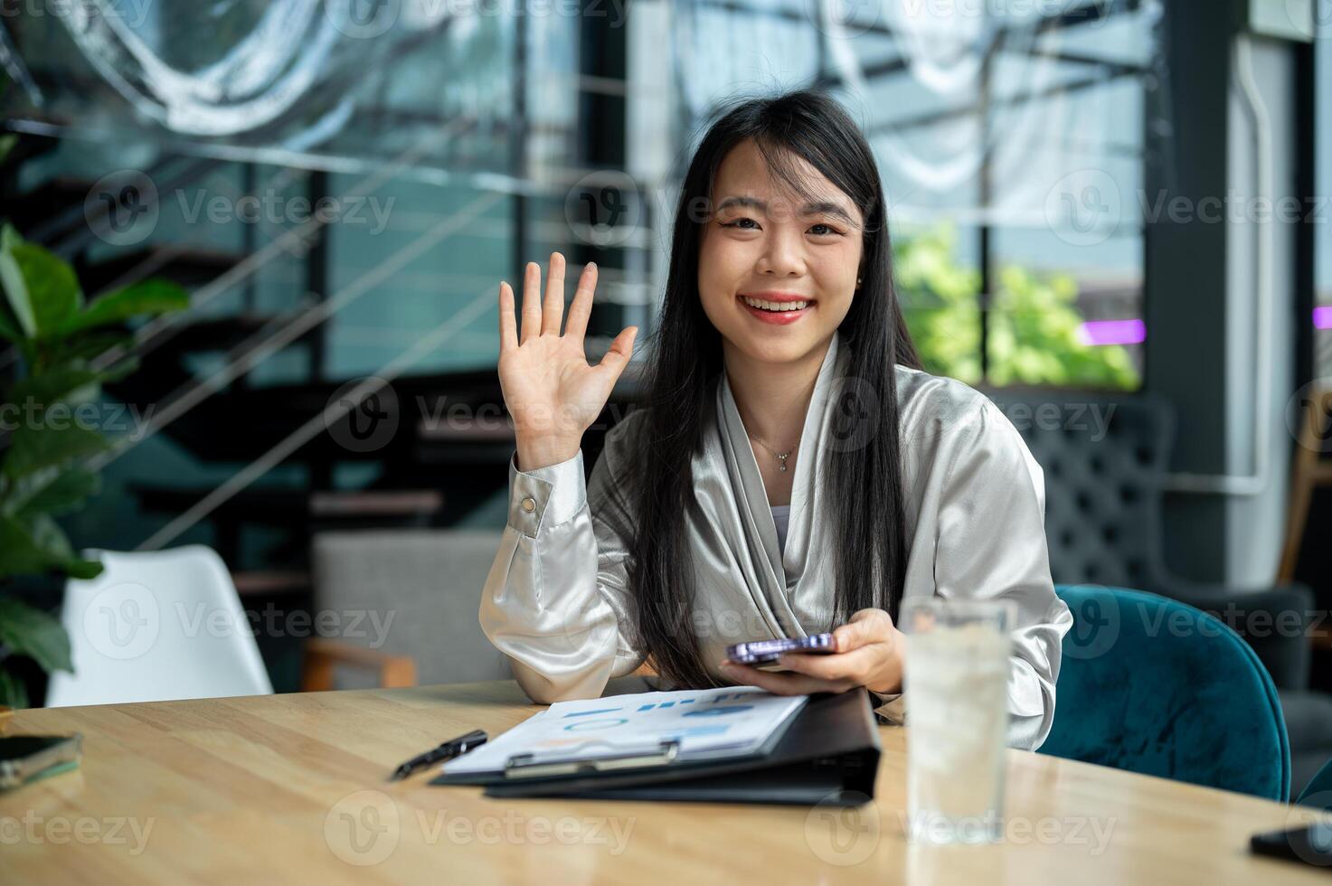A confident Asian businesswoman is waving her hand and smiling at the camera while sitting at a desk photo