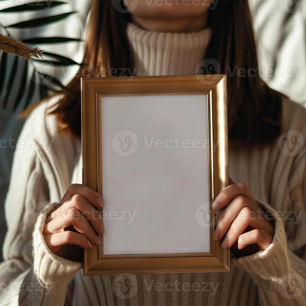 Woman in a cozy sweater holding a blank picture frame, ready for customization. Warm lighting and soft textures create a welcoming atmosphere. photo