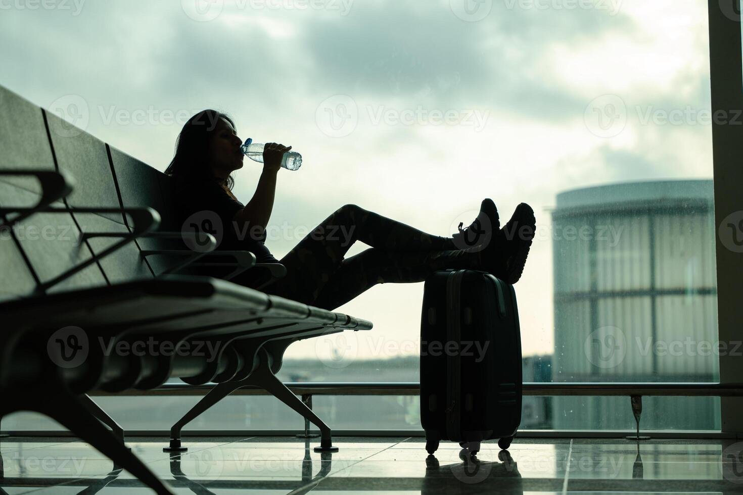 Woman sitting on an airport bench, drinking a bottle of water while waiting for her flight, with a background of sky with clouds and low angle shot photo