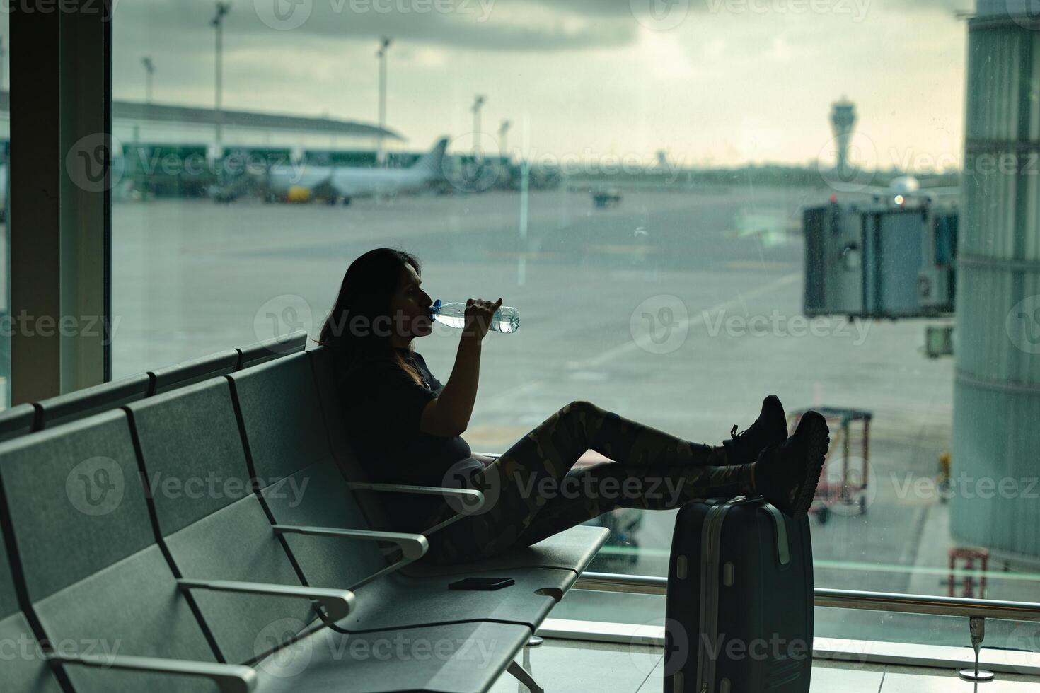 Woman drinking a bottle of water while waiting for her flight sitting and relaxing on a bench at the airport. photo