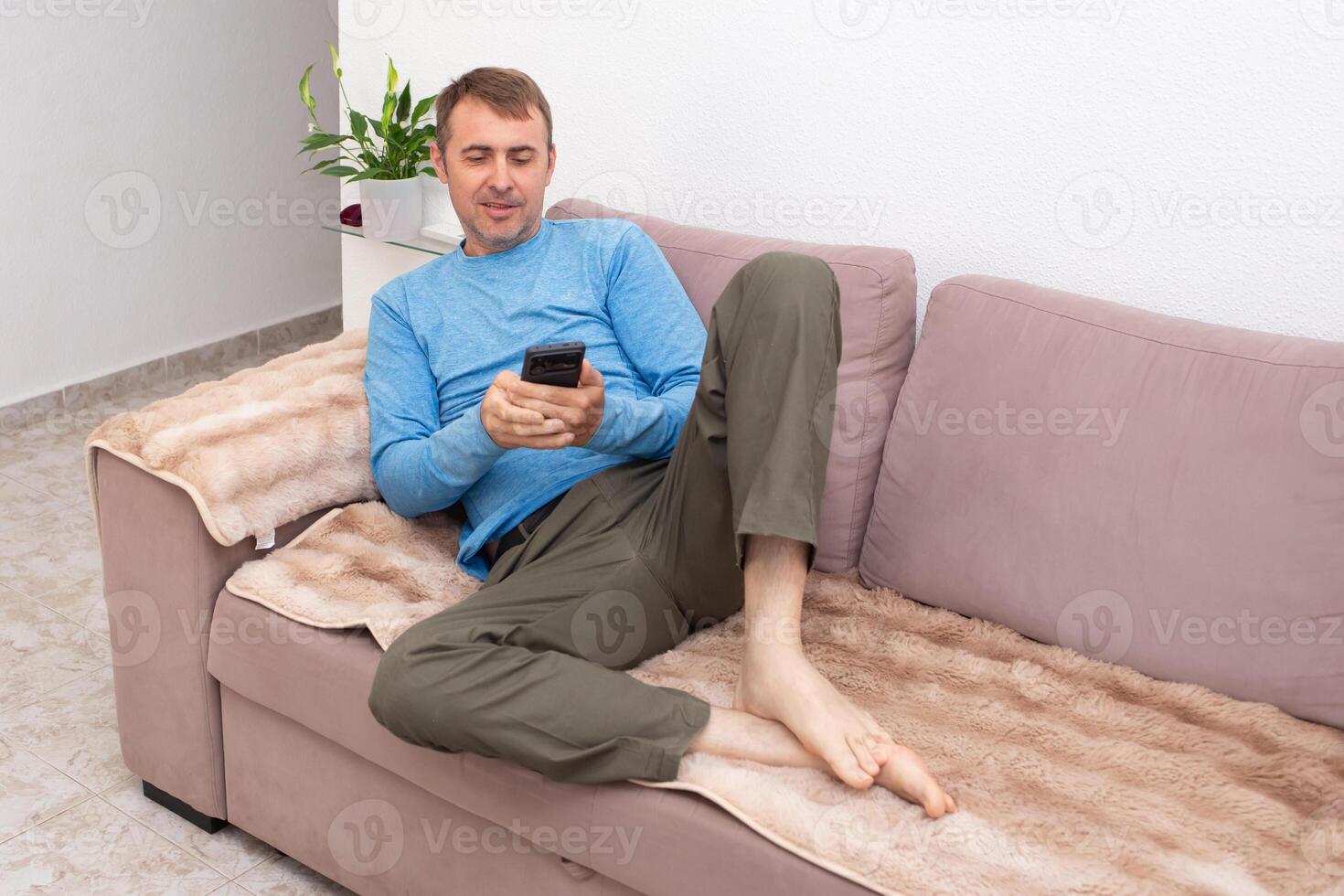 A cheerful man is texting his friends and agreeing on a meeting place while sitting on the sofa in his living room photo