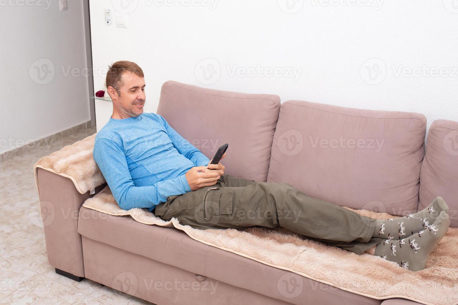 A cheerful man is texting his friends and agreeing on a meeting place while sitting on the sofa in his living room photo