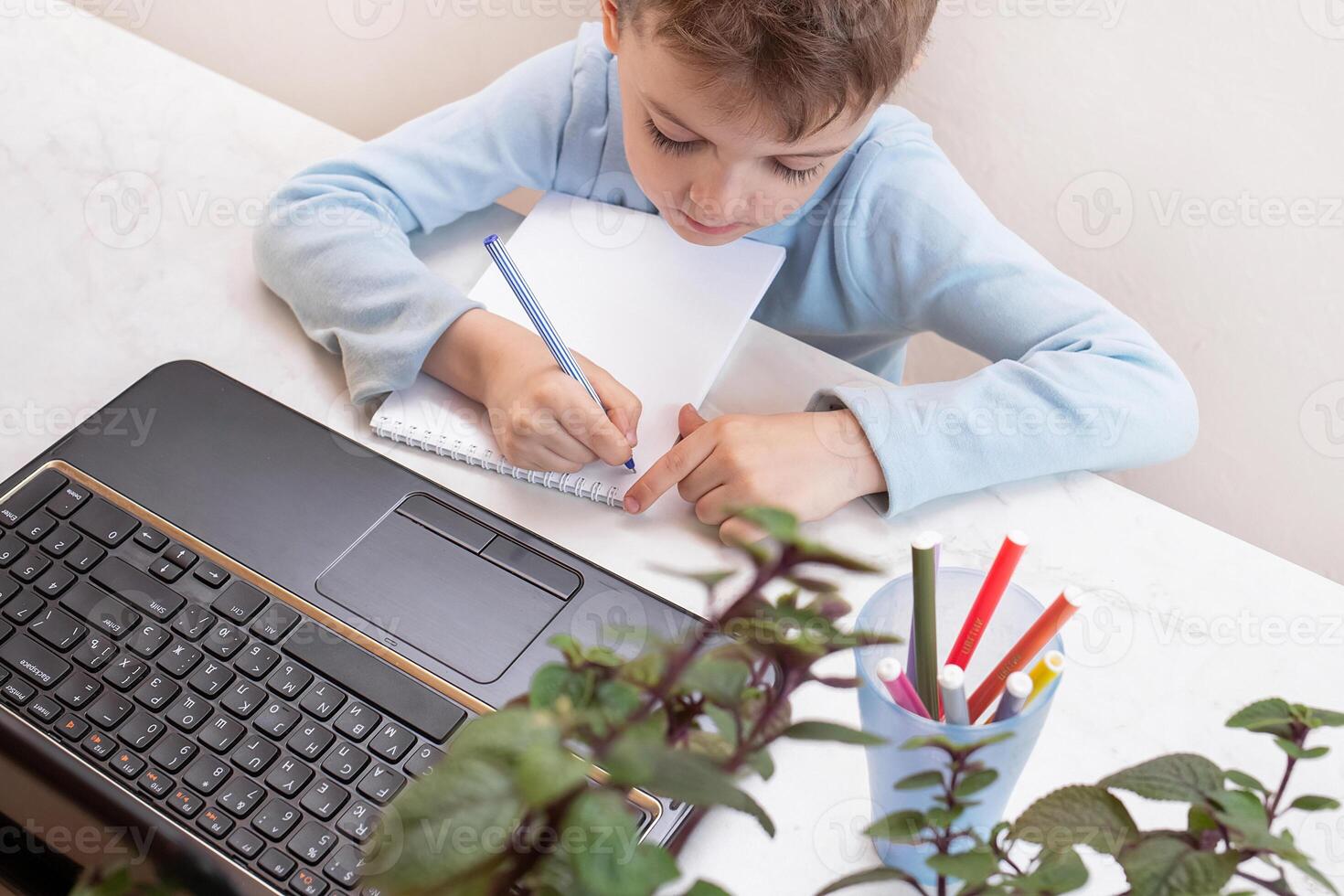 close-up top view, a serious child in a blue sweater writes in a notepad during an online lesson. teaching people at home in bedroom, studying online on computer photo
