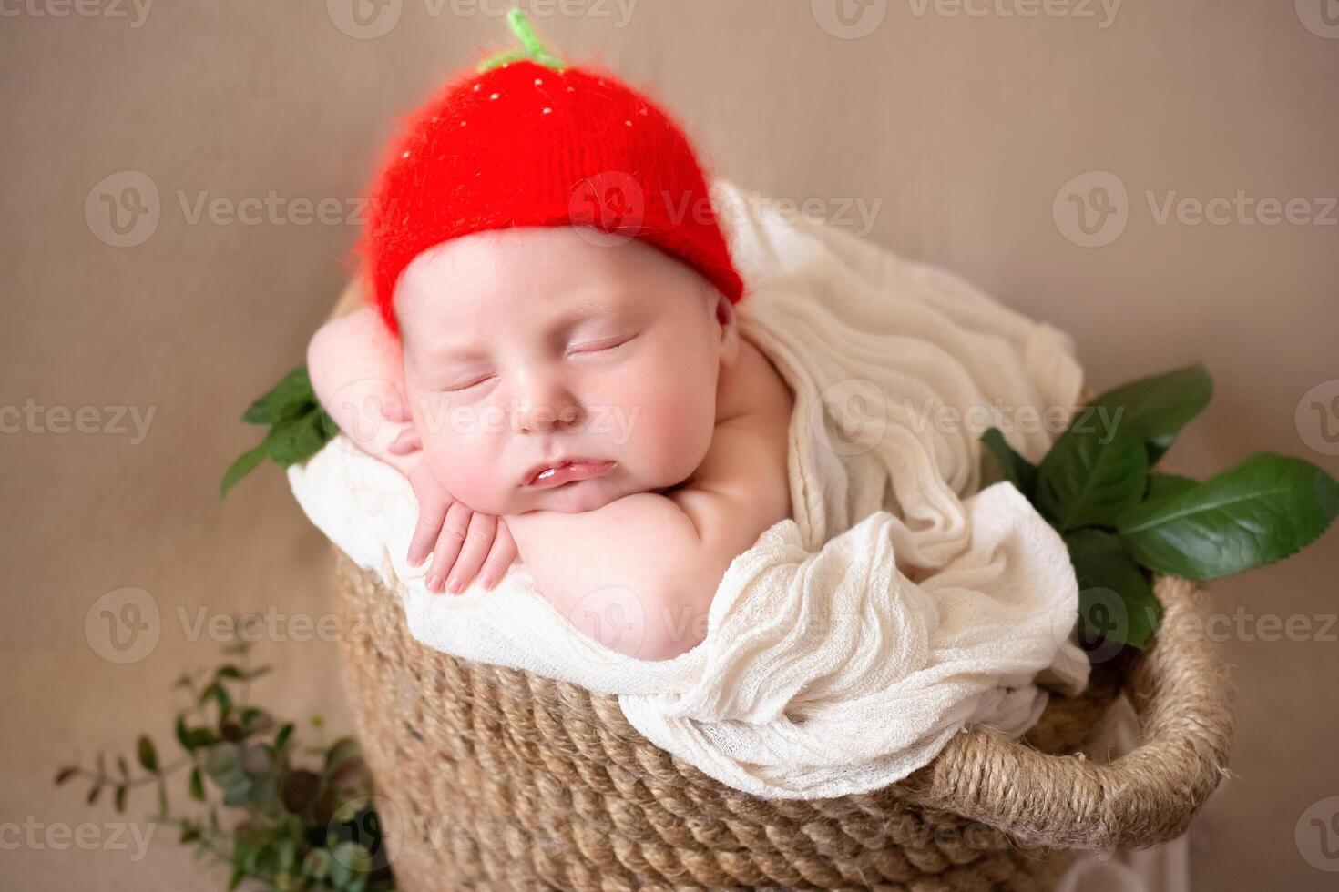 a cute newborn iin a hat of strawberries in the form of a sunflower flower sleeps peacefully. beige background with space for text with a baby sleeping in a wicker basket decorated with green leaves. photo