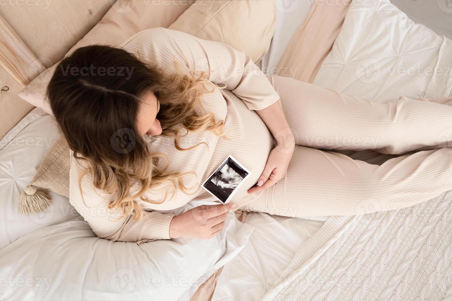 a beautiful pregnant woman with long blond hair has breakfast in a large bright bedroom on a bed covered with a white blanket. boho style. view from above photo