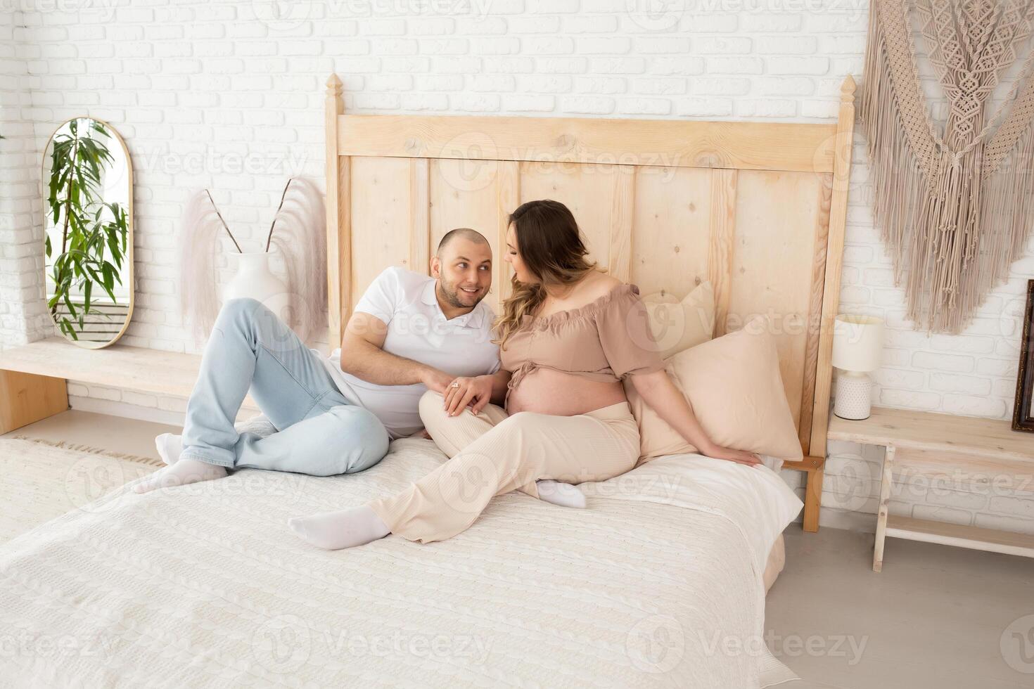 husband and pregnant wife are sitting and talking on a bed covered with a white knitted bedspread. family in a beautiful bright bedroom with large windows photo
