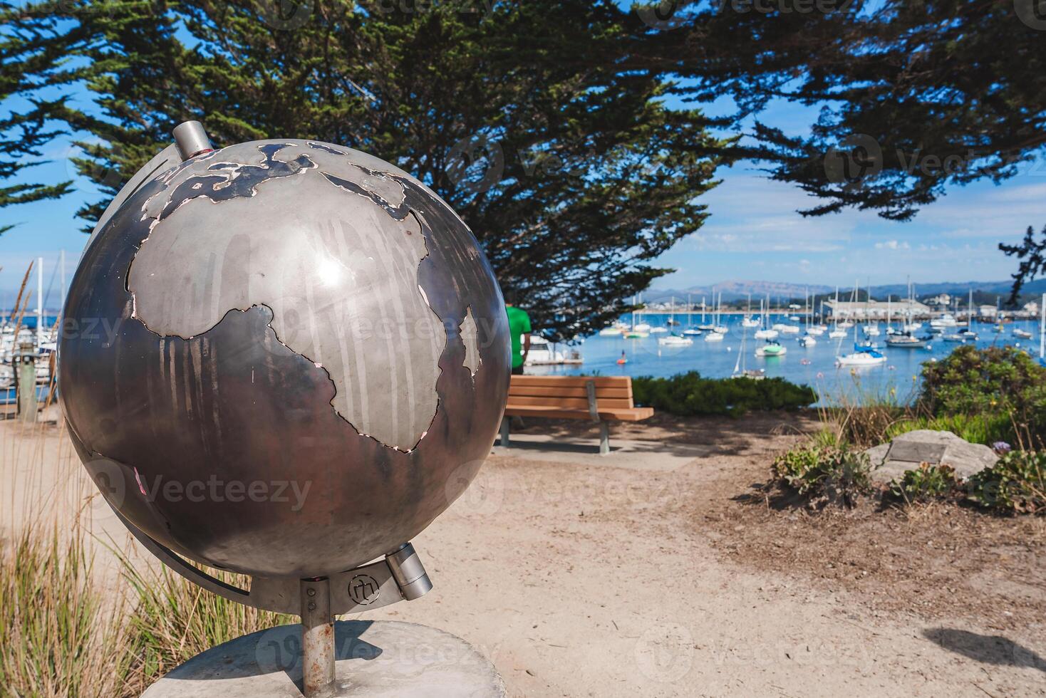 Metallic globe sculpture on stand with reflective surface featuring continents outlined against clear blue sky. Harbor view with boats and lush greenery in background. Peaceful coastal setting. photo