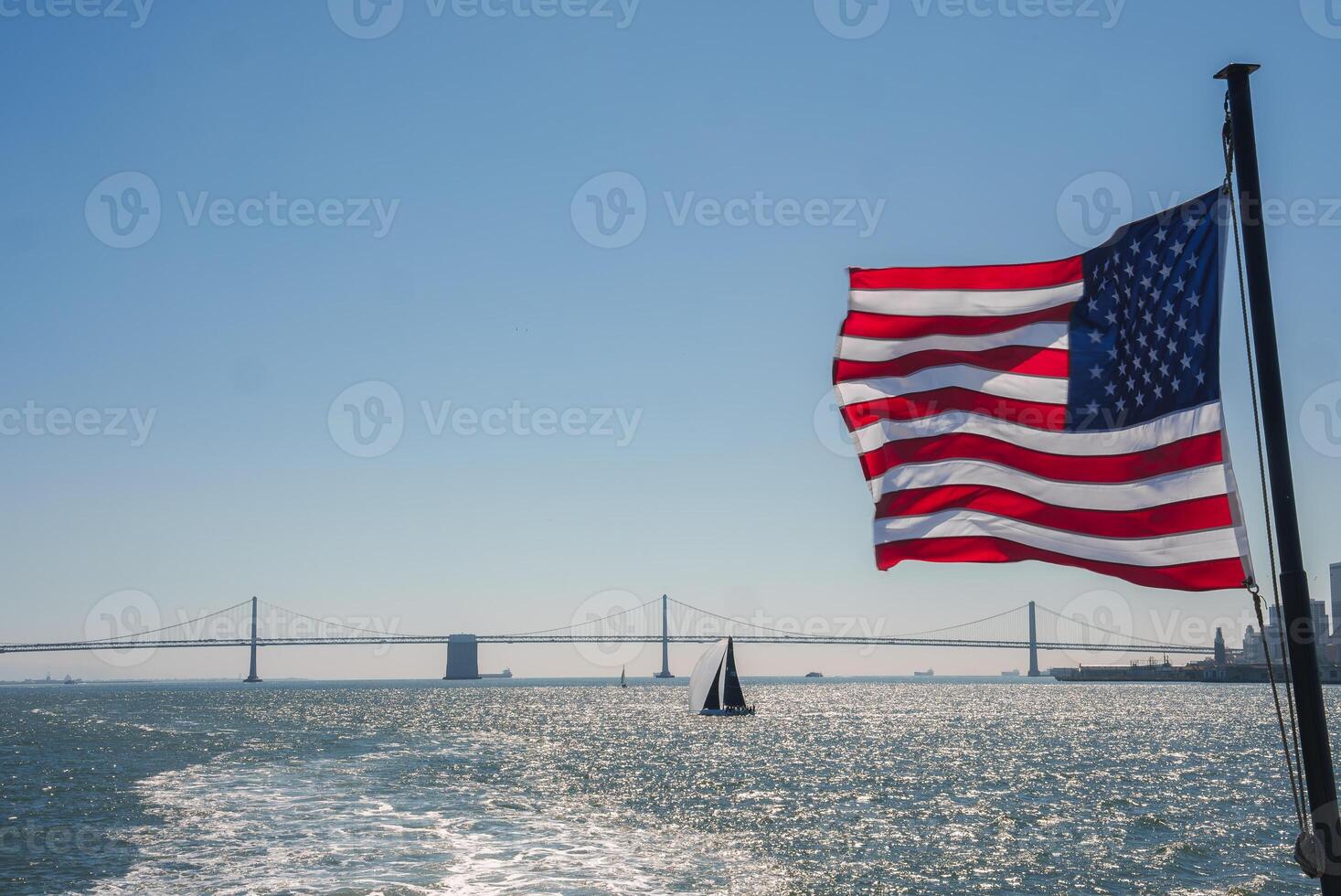 American flag waving in the wind with the Golden Gate Bridge in the background, overlooking San Francisco's cityscape. Tranquil scene with sailboat passing by. photo