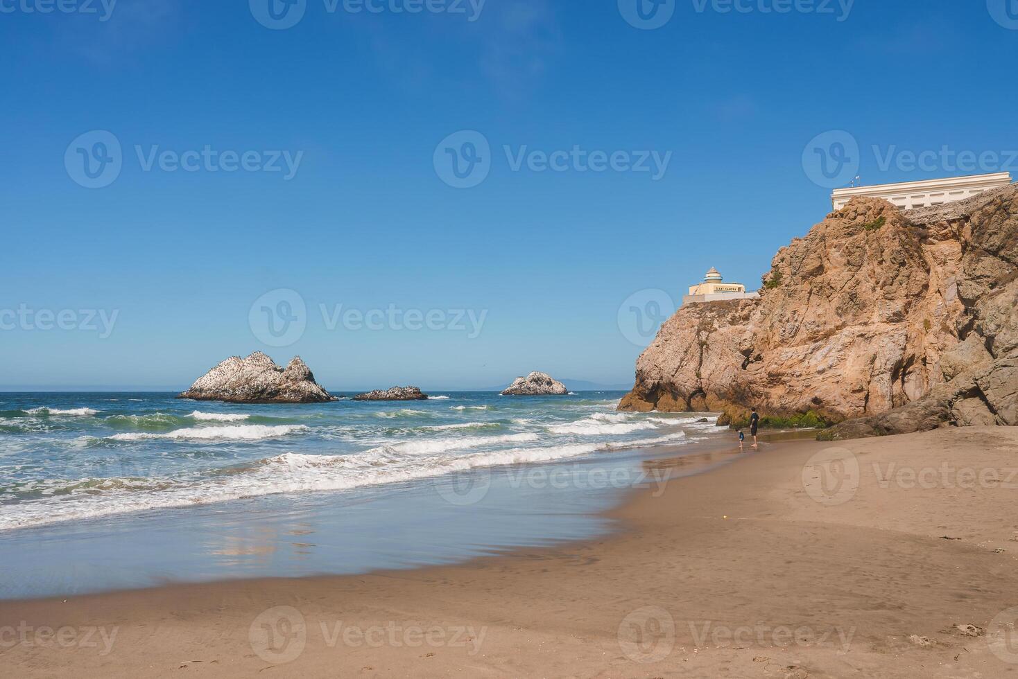 Tranquil San Francisco beach scene with sandy shore, lapping waves, rocks, and a cliff with a building on top. Peaceful ambiance with few individuals. photo