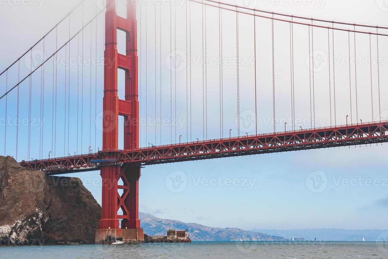 Iconic Golden Gate Bridge in San Francisco, California. View from below shows red orange structure, symmetrical cables, rocky outcrop, and distant land. photo