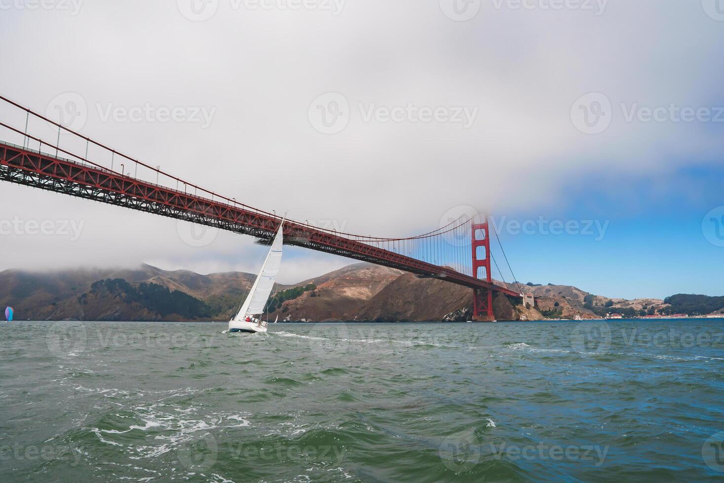 Serene maritime scene in San Francisco with the Golden Gate Bridge in the backdrop. White sailboat, partly cloudy sky, and misty hills add natural mystique. photo