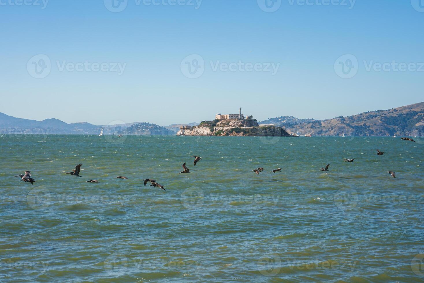 View of Alcatraz Island in San Francisco Bay, USA. Alcatraz prison atop the rocky outcrop visible against clear blue sky. Birds in choppy waters, hills in the distance. Serene sunny day scene. photo