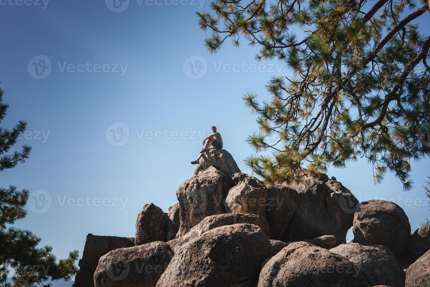 Person sitting on large boulders, wearing summer clothes, gazing into the distance under a clear blue sky. Pine tree in background. Serene outdoor scene. photo