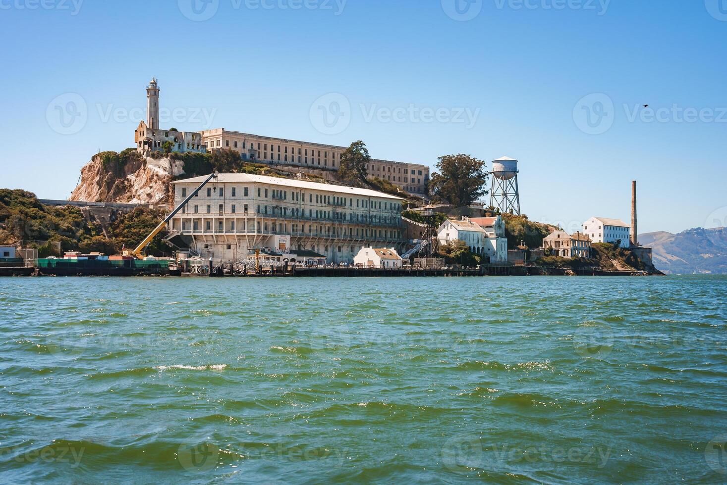 Clear, sunny view of Alcatraz Island in San Francisco Bay. Photo from the water, showing former prison complex and iconic lighthouse. Rocks, buildings, and blue sky in background.