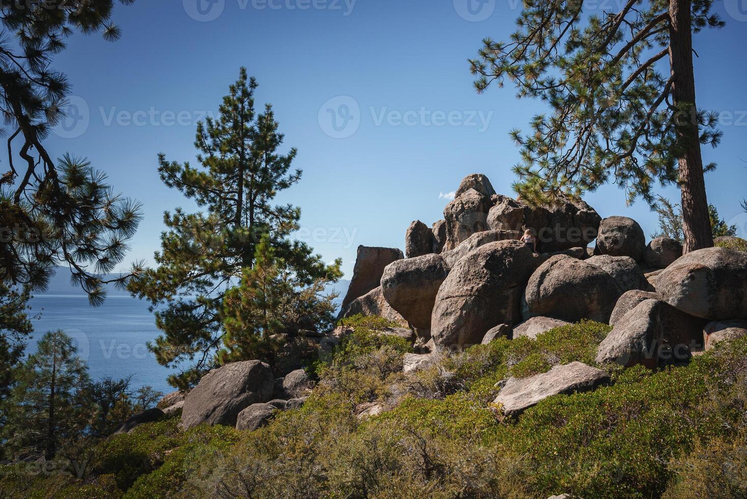 Serene natural landscape with rocky outcrop, lush greenery, tall pine trees, and person sitting on boulder. Tranquil scene near calm body of water. Mountainous region vibes in Tahoe. photo
