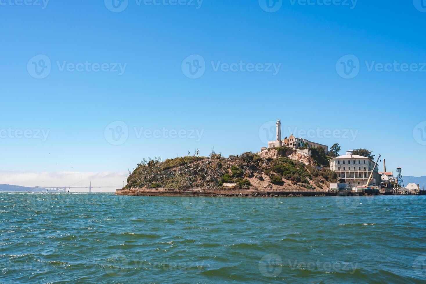 Clear view of Alcatraz Island in San Francisco Bay, California, USA. Features the prison facility, lighthouse, and rugged terrain. Golden Gate Bridge in the background. Sky is clear over calm waters. photo