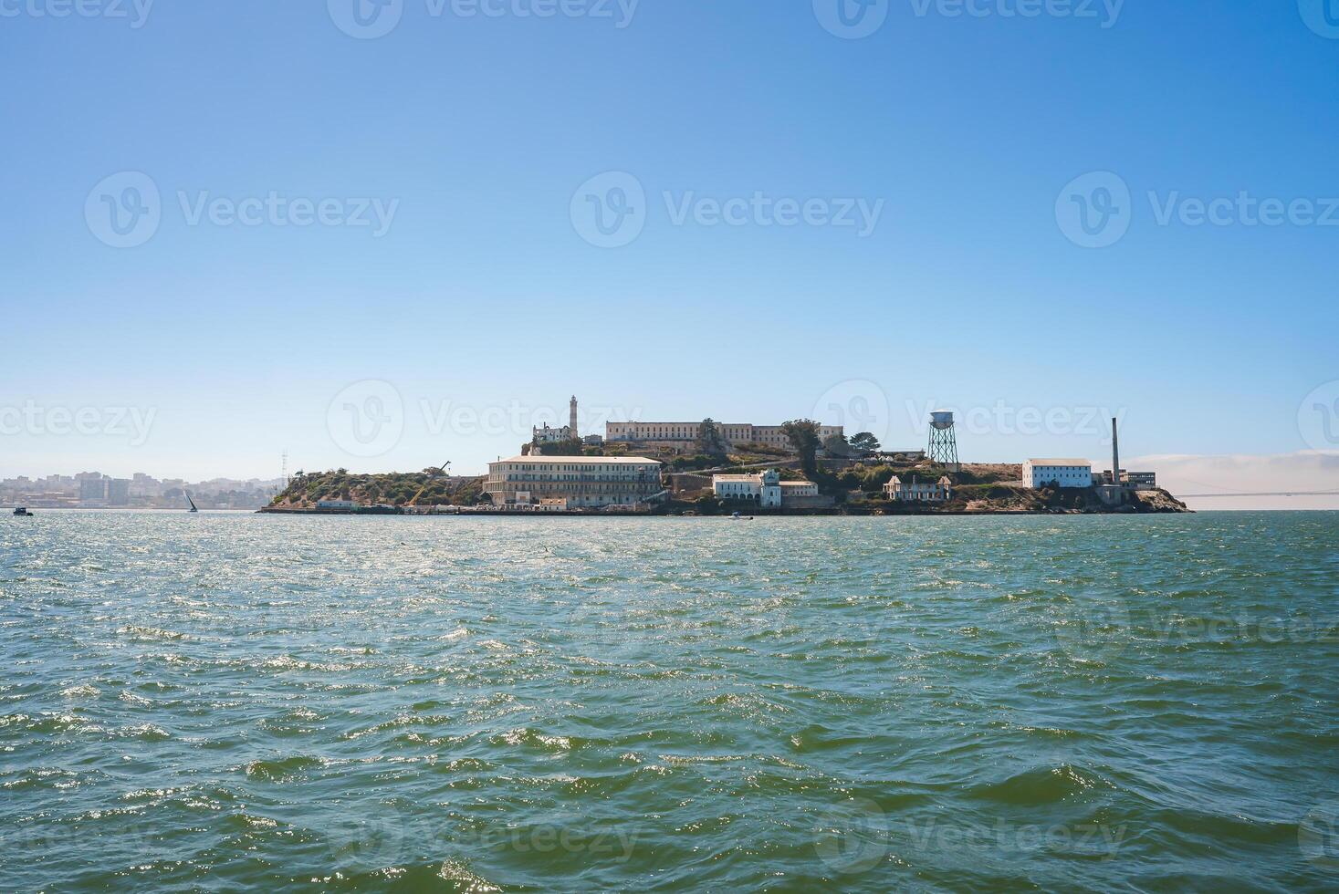 Scenic view of Alcatraz Island in San Francisco Bay on a sunny day. Shows the former prison complex with main cellhouse, water tower, and skyline in the distance. photo