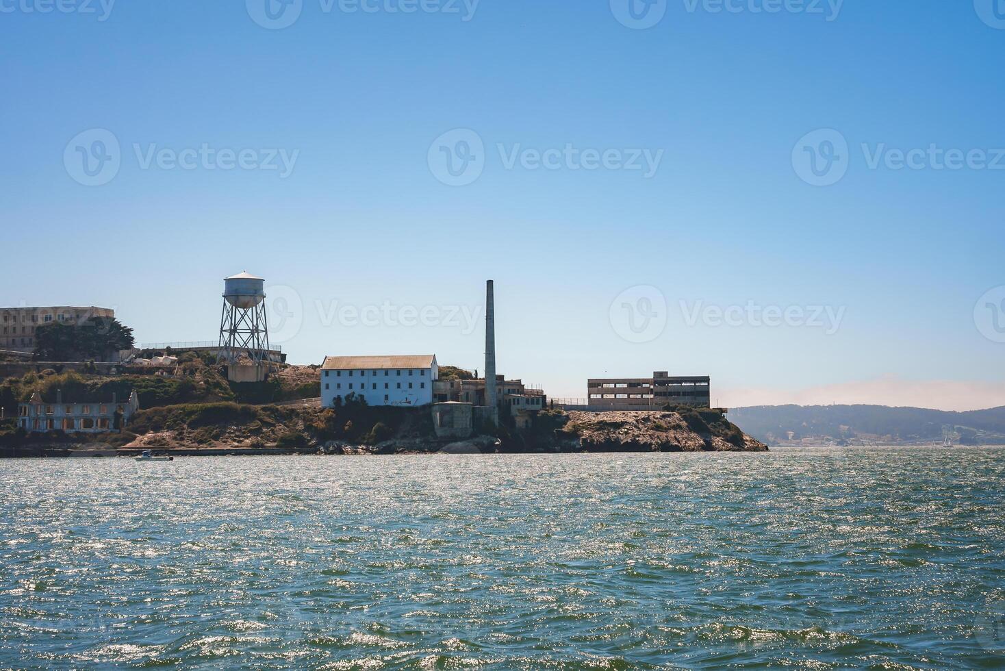 View of Alcatraz Island on a clear day in San Francisco Bay. Features water tower, buildings, smokestack, and boat, capturing former prison's history. photo