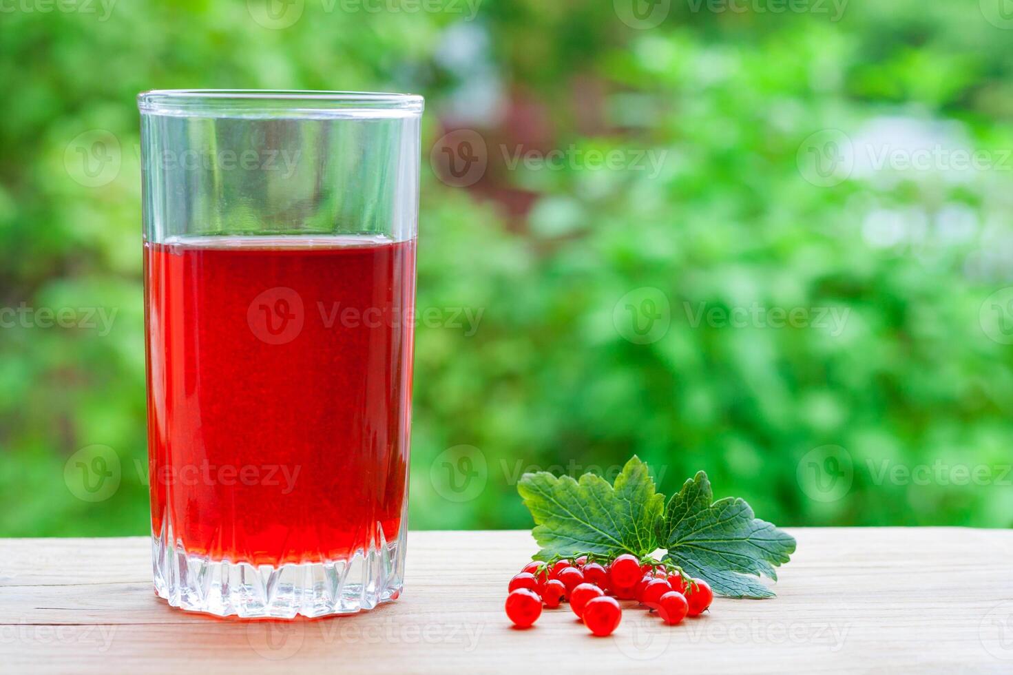 A glass with red juice and branch of redcurrant with leaves on a wooden table on a background of green garden in blur with copy space photo