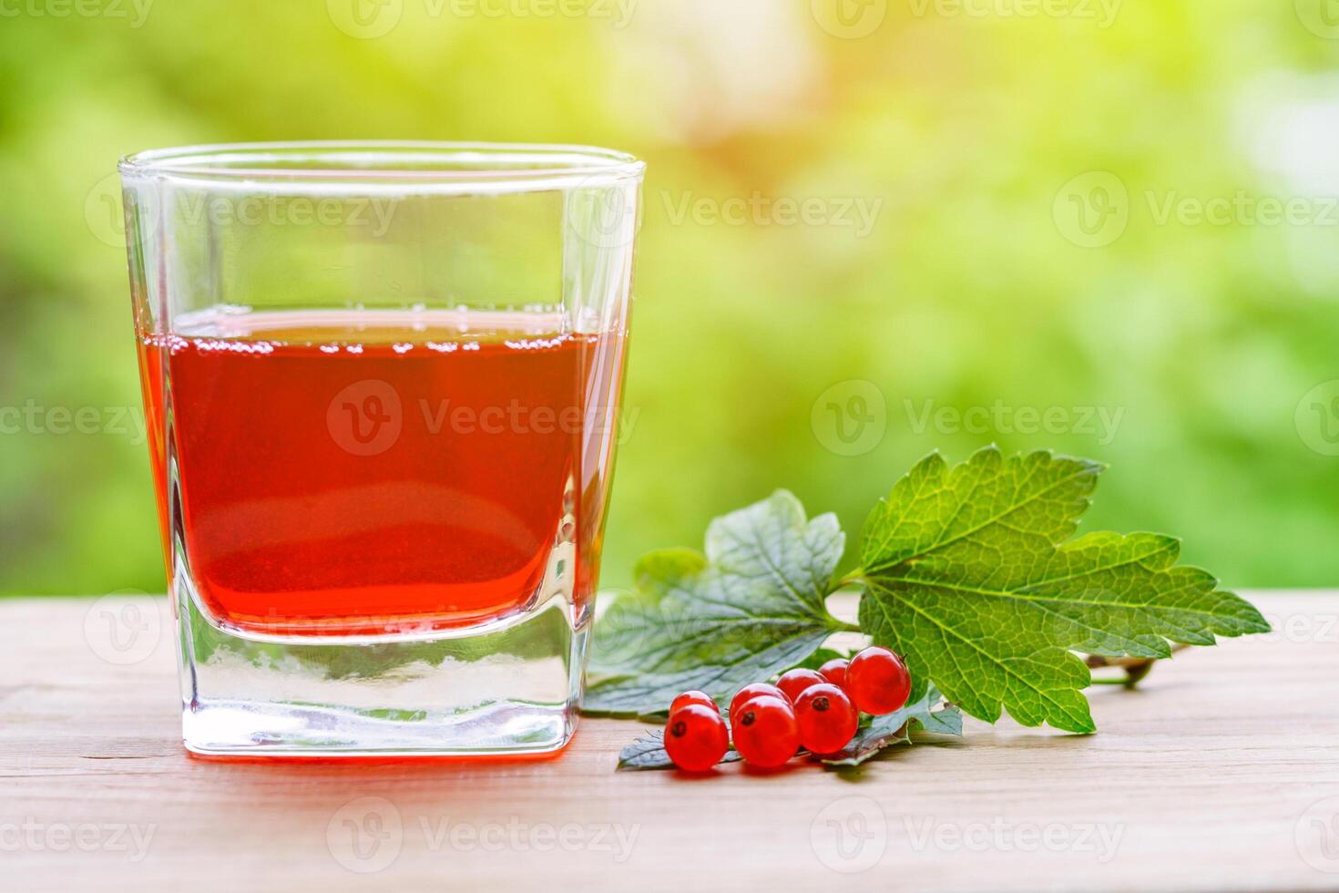 A glass with red currant juice and a branch of berries with leaves on a wooden table, illuminated by sunlight, on a background of green garden in blur with copy space photo