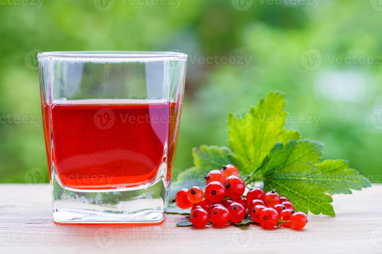 View on a glass with red juice and a branch of redcurrant berries with leaves on a wooden board on a background of green garden in bokeh. Summer concept photo