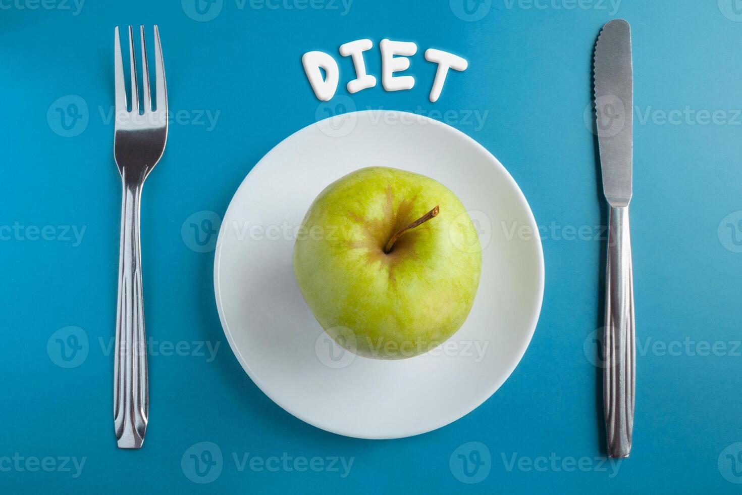 Top view of a green apple on a saucer, with a fork and knife on a blue background with an inscription diet. Concept photo