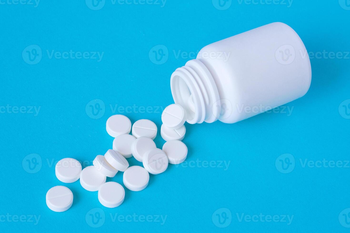 A pile of white tablets poured from a white plastic jar on a blue table photo