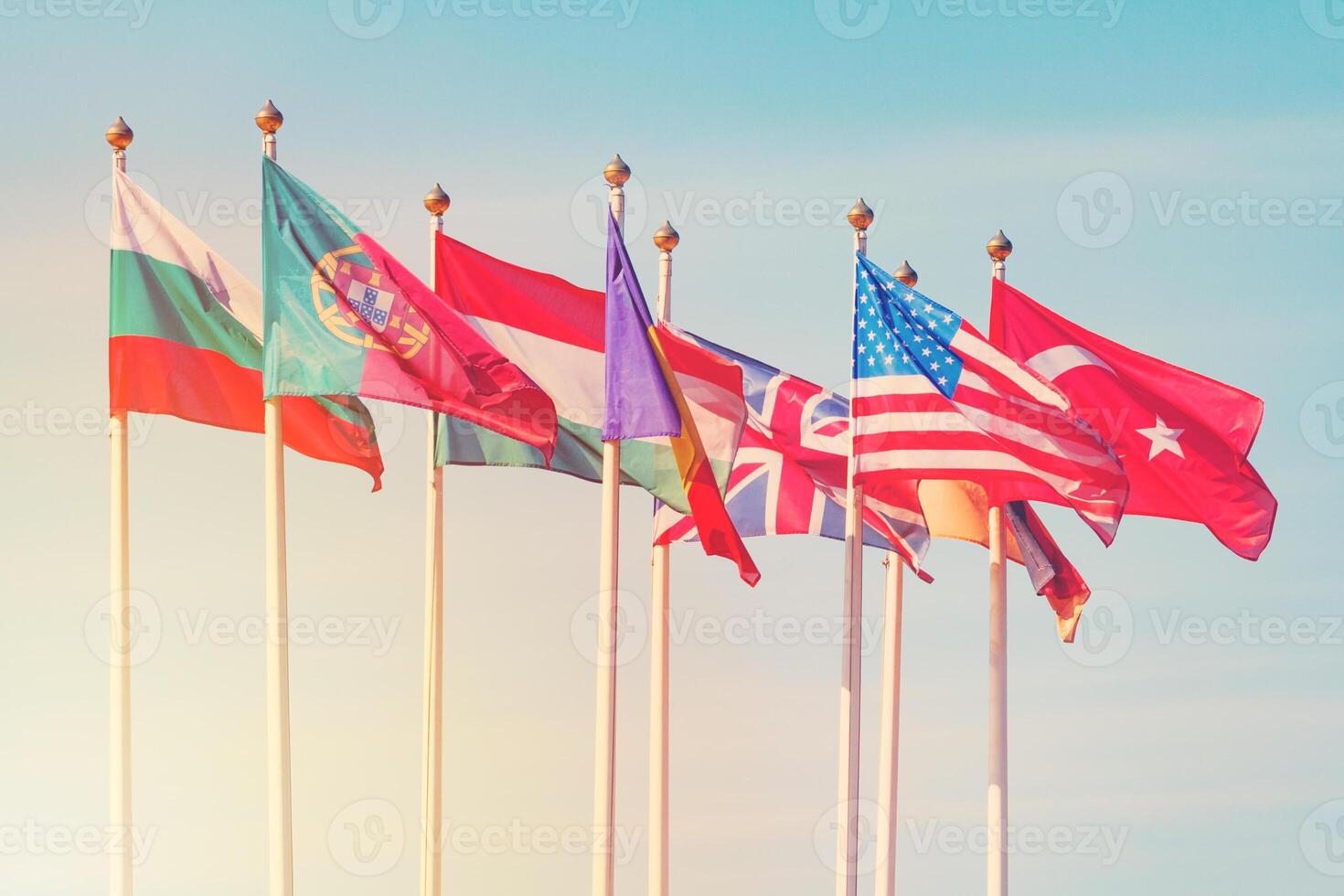 Flags of different countries on the white flagpoles flutters in the wind against a sunlight photo