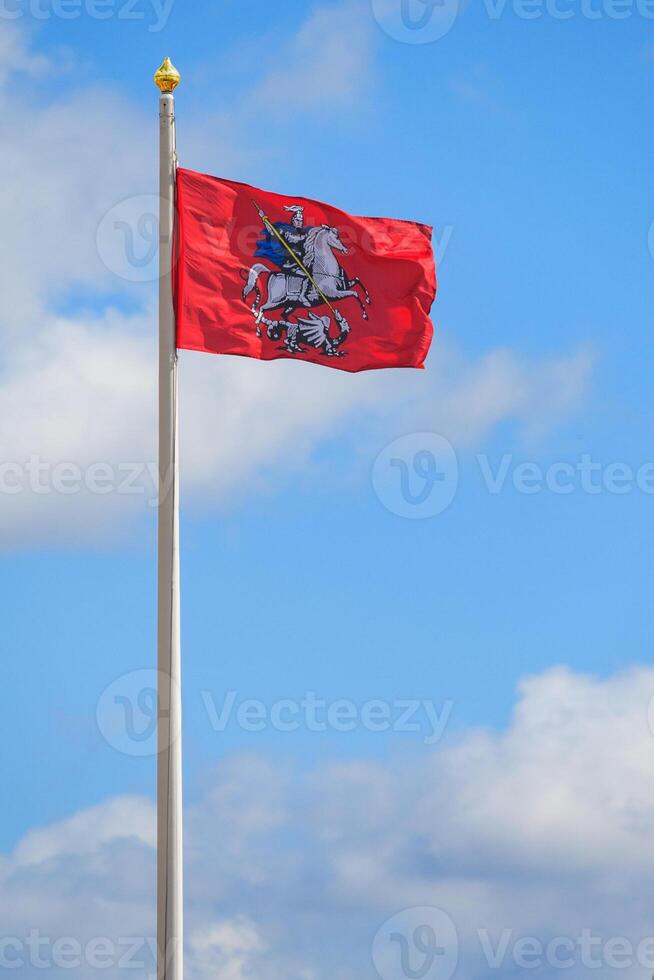 The red flag of the city of Moscow with the coat of arms - St. George the Victorious on horseback, will strike the Serpent with a spear, against a blue sky with clouds photo