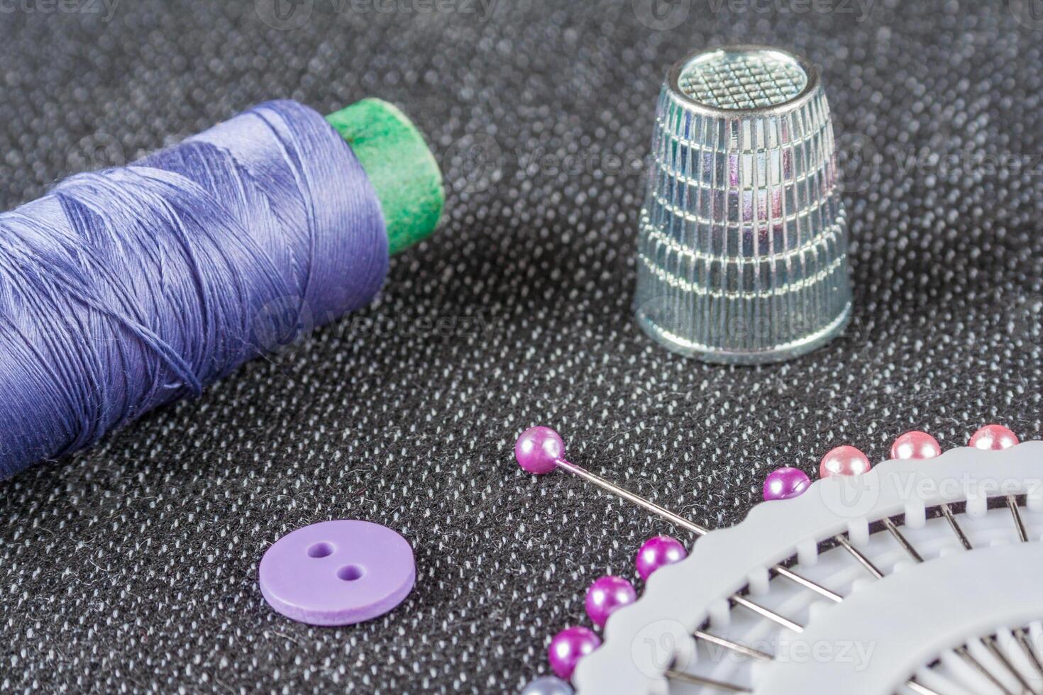 Sewing accessories violet spool of thread, needles, thimble, button on the textured fabric surface photo