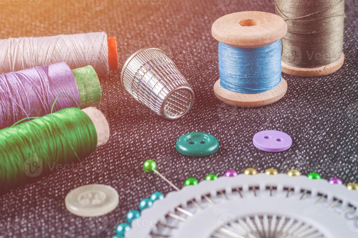 Sewing accessories spools of threads, needles, thimble, buttons, on the textured fabric surface illuminated by warm light photo