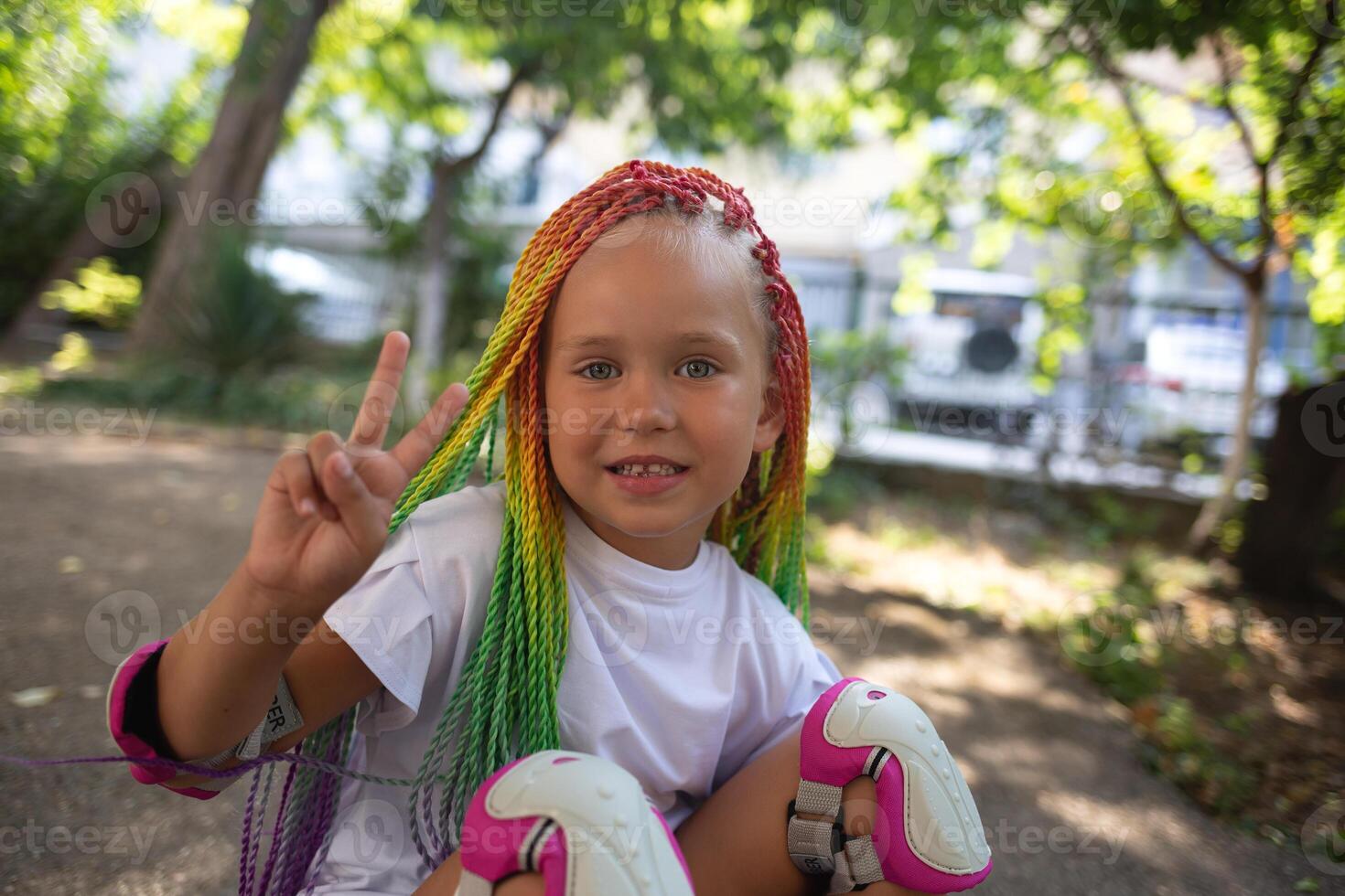 little cute girl with colorful afro pigtails sits on a skateboard and shows a peace sign photo