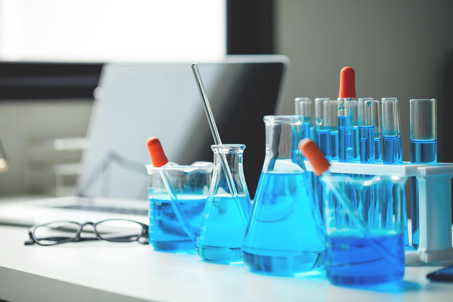 A table with a laptop, a few beakers and test tubes, and a blue liquid. The table is set up for a science experiment photo