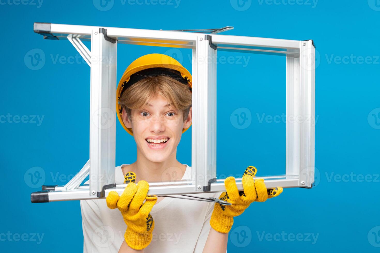 Happy repairman teen in white t-shirt and gloves with yellow helmet in head, holding ladder in hands standing in studio on blue background. A future architect isolated on a blue background photo