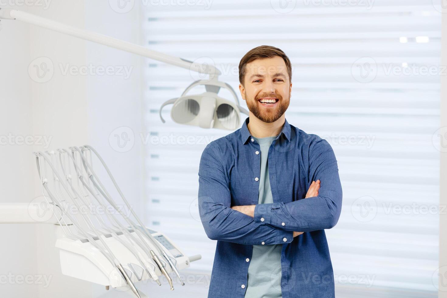 Confident male patient of stomatology standing in modern clinic. Man smiling at the camera crossing his arms in front of him photo