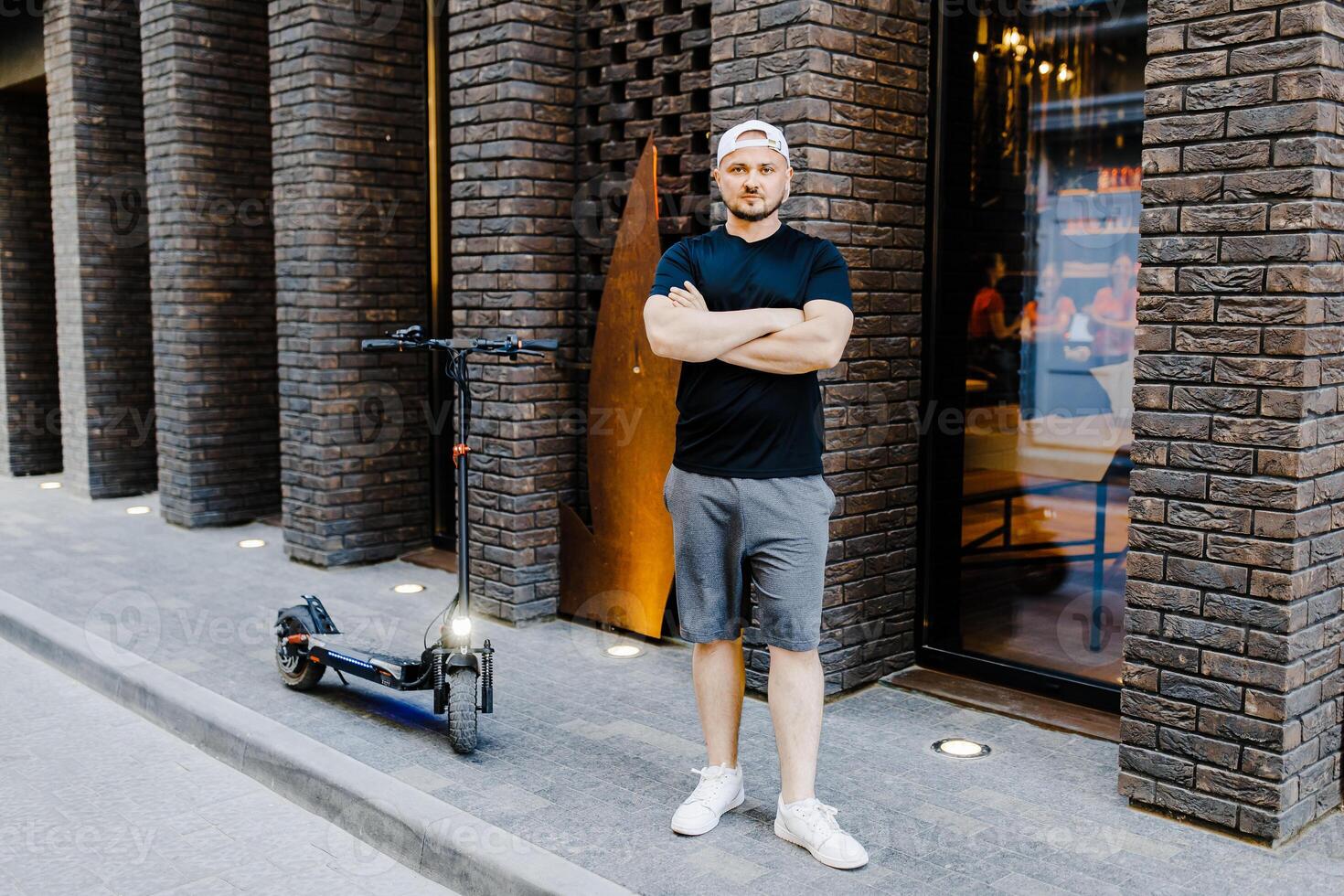 Young handsome man in n white baseball cap and casual clothies standing near electric scooter on the street. photo