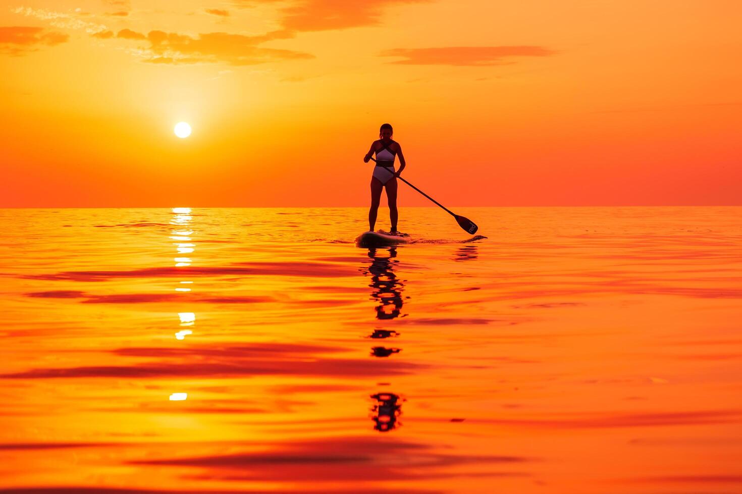 June 25, 2021. Anapa, Russia. Silhouette of woman paddle on stand up paddle board at quiet sea with sunset. Woman on Red Paddle sup board and sunset with reflection photo