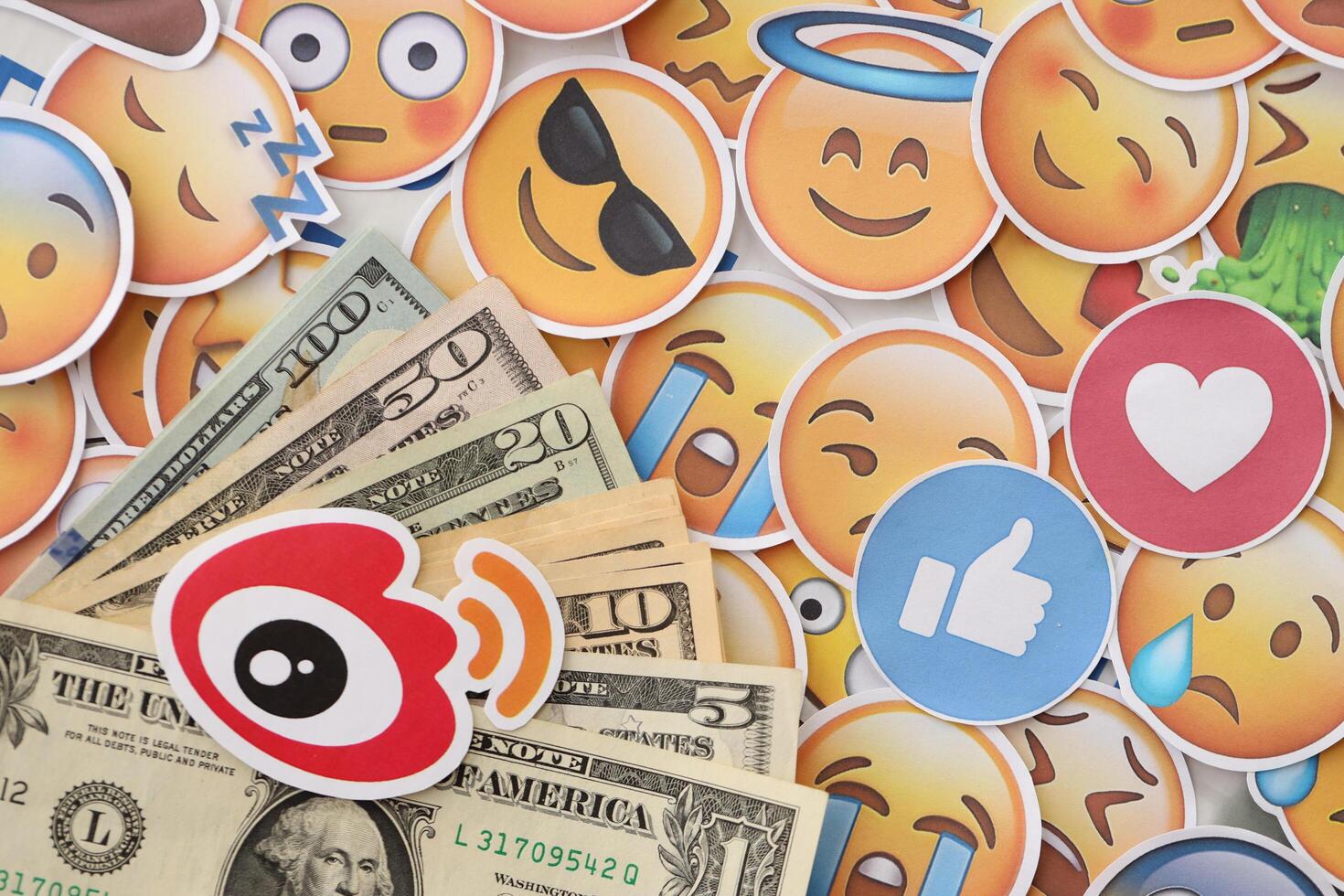 KYIV, UKRAINE - APRIL 20, 2024 Weibo logo and many usd money bills. Symbol of famous social network platform and dollar currency banknotes on lot of emoji reactions photo