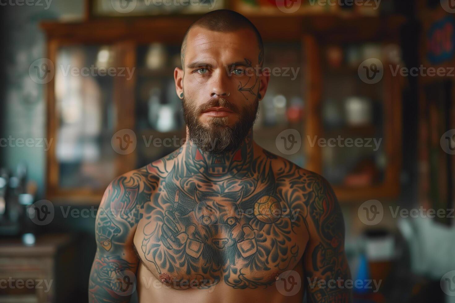 Bearded man with chest and arm tattoos posing in front of a wooden cabinet photo
