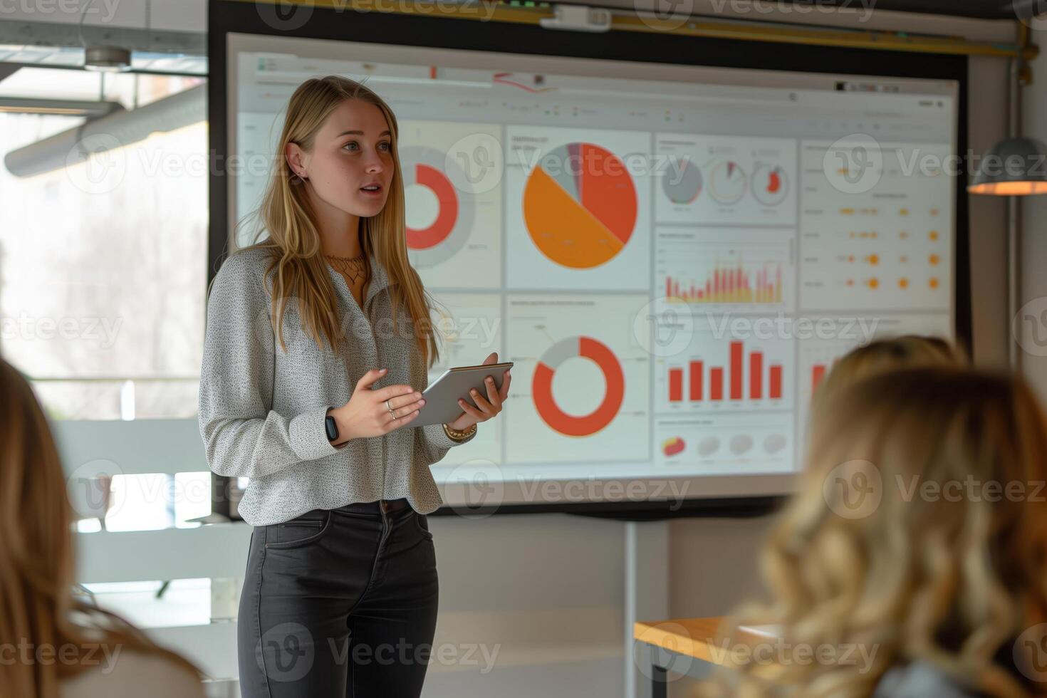 A businesswoman is presenting data charts to her colleagues during a meeting. She is holding a tablet and standing in front of a large screen displaying graphs and charts photo