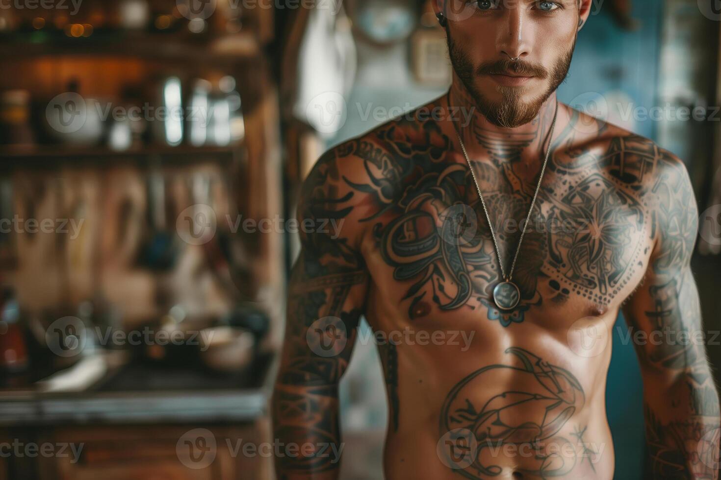 man with a beard and numerous tattoos standing shirtless in an interior space. The man has a muscular physique and a chain necklace photo