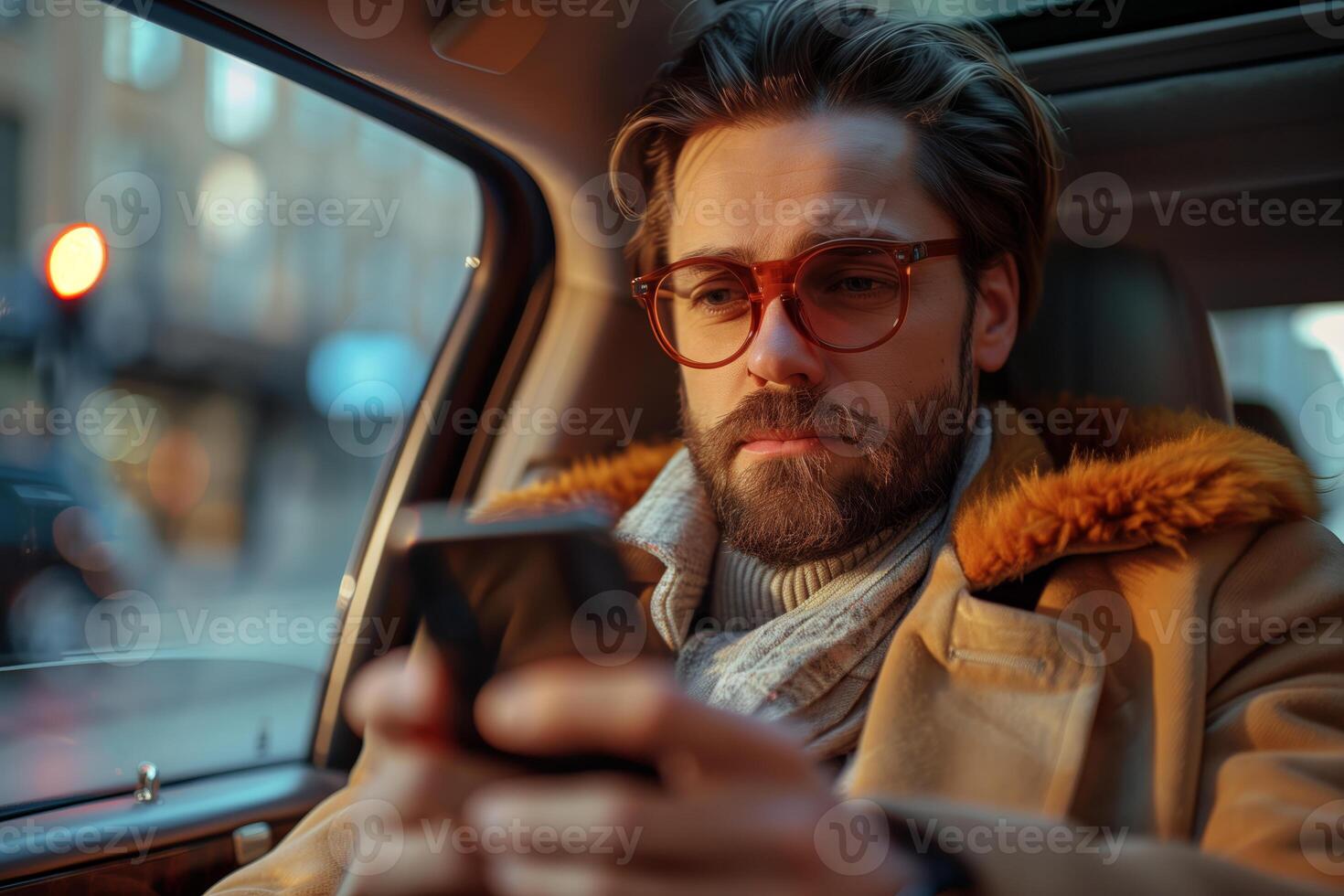 A man with a beard and glasses is sitting in the passenger seat of a car, using his smartphone. He is wearing a brown coat with a fur collar and a white turtleneck sweater photo