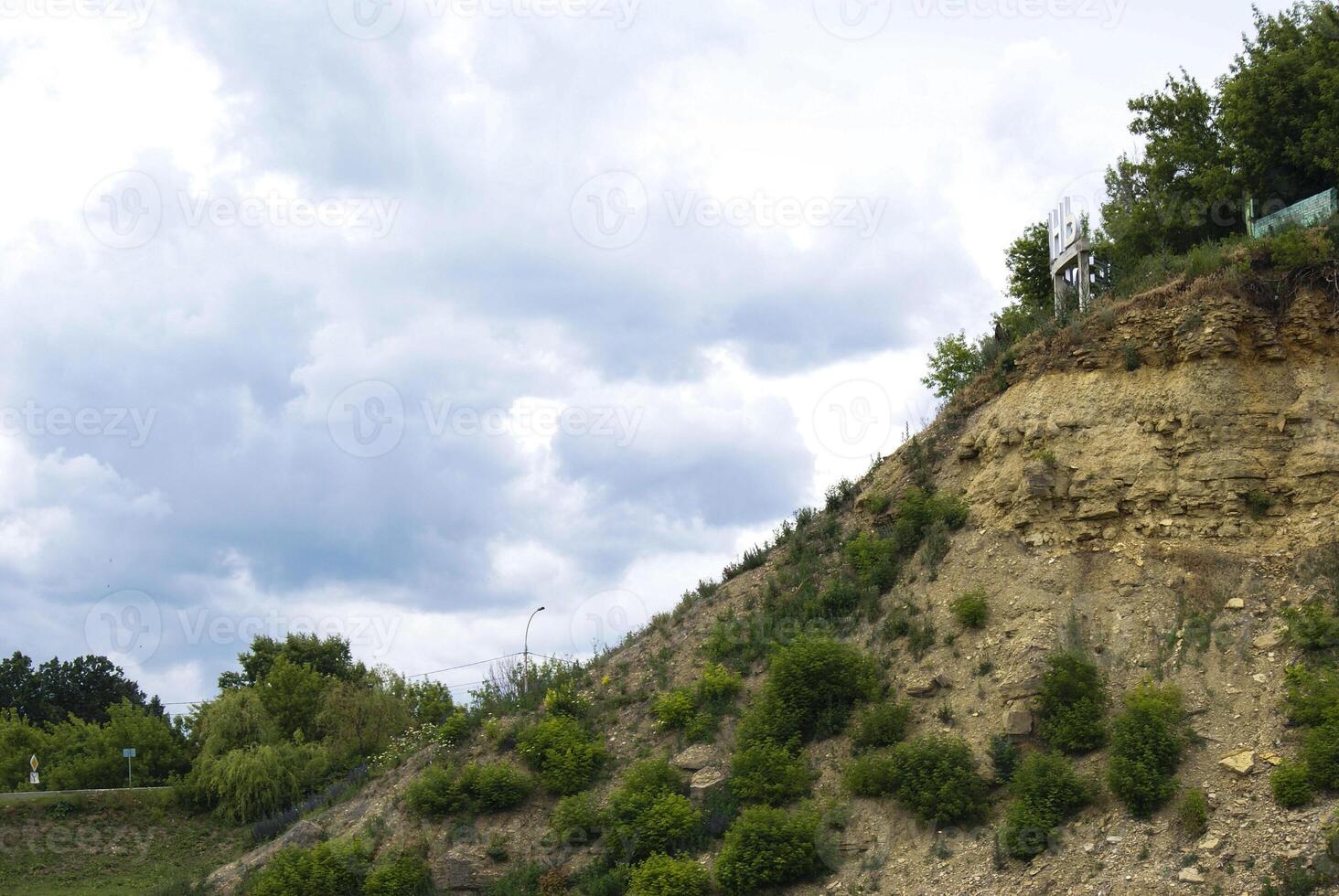 A hillside with a rocky, layered slope and green vegetation. The sky above is filled with white, puffy clouds. photo
