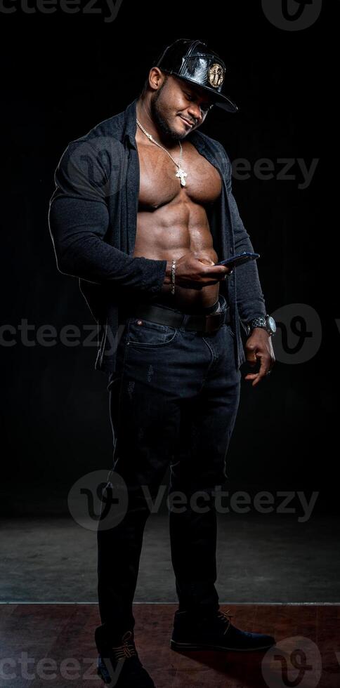 Strong athlete in dark jeans poses to the camera on black background. Unbuttoned black shirt and perfect abs. Portrait of an afro american bodybuilder with phone in hands. photo