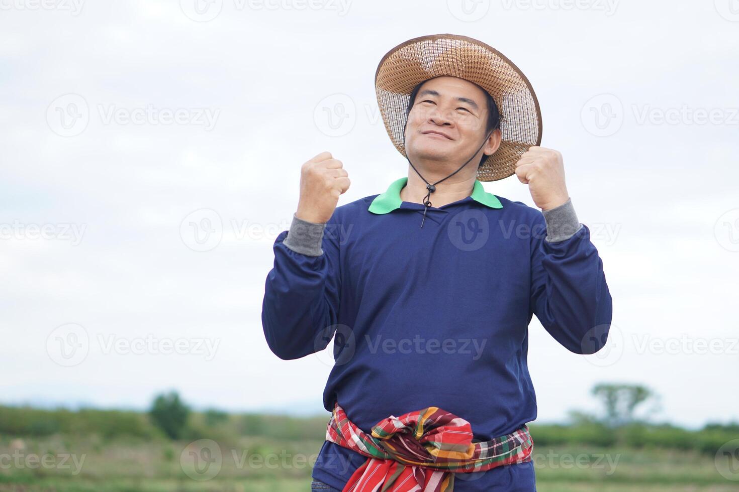 Handsome Asian man farmer stands at agricultural land, wears hat, Thai loincloth on waist, raises fists up with excited feeling or success. Concept, agriculture occupation. photo