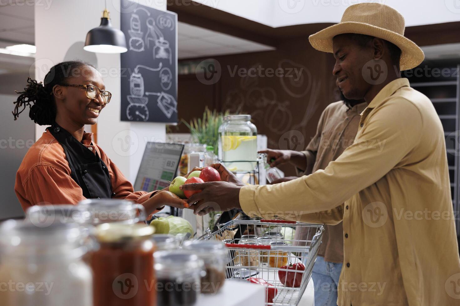 African American shopkeeper of an eco friendly business with fresh organic items helping clients. Focus on a black man presenting fruits to a female employee at the checkout desk. side view shot. photo
